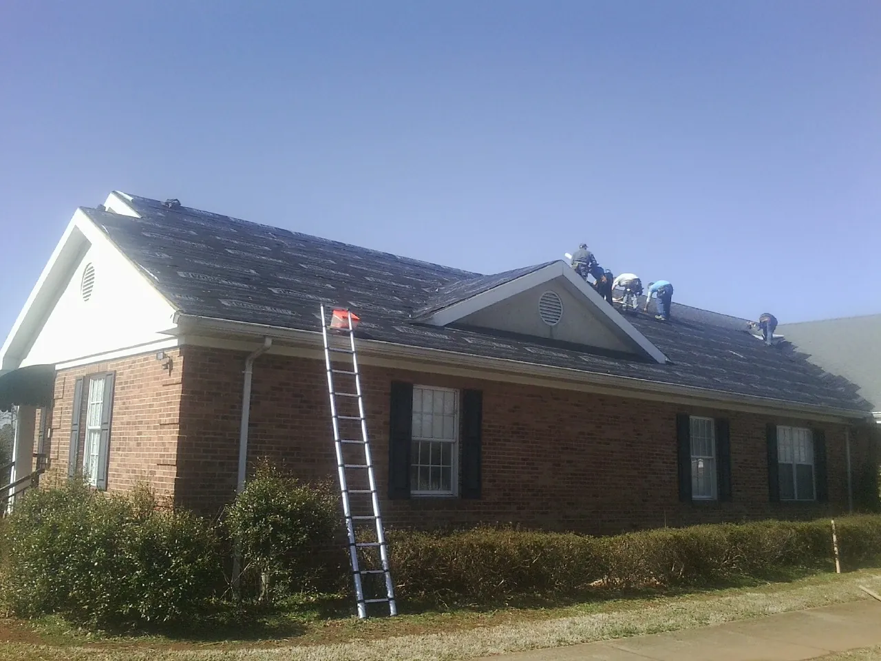 Crew tearing shingles off a brick ranch roof with a ladder set — Emergency Roof Repair work by our crew in Vardaman