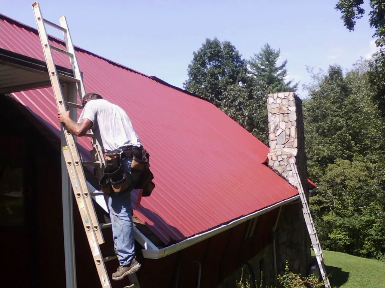 Roofer on a ladder beside a red metal roof and stone chimney — Soffit & Fascia Repair by Roofing Brothers in Vardaman